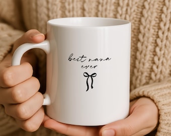 Op de afbeelding: Een witte keramische mok met de zwarte tekst "best nana ever" in een cursief lettertype, geplaatst boven een eenvoudige zwarte lijntekening van een gestrikte lintstrik. De mok heeft een gladde afwerking en een standaard gebogen handvat.