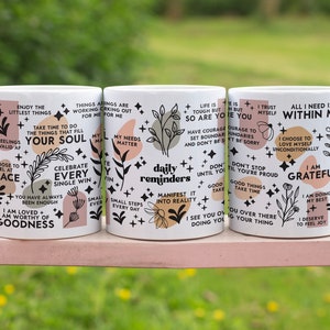 Puede incluir: Tres tazas de cer&aacute;mica blanca con dise&ntilde;os florales negros y rosas y citas inspiradoras. Las citas incluyen "Disfruta de las peque&ntilde;as cosas", "Celebra cada victoria", "Recordatorios diarios", "La vida es dura pero t&uacute; tambi&eacute;n", "Todo lo que necesito est&aacute; dentro de m&iacute;", "Estoy agradecido".