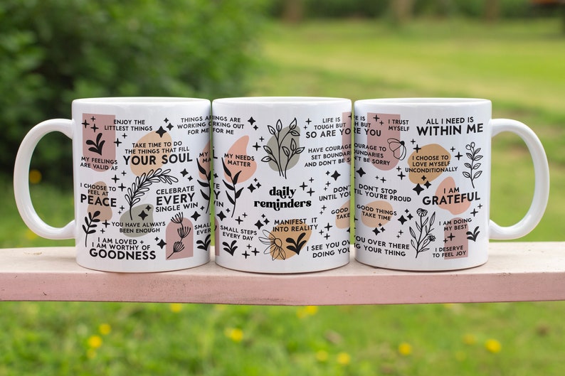 Puede incluir: Tres tazas de cer&aacute;mica blanca con dise&ntilde;os florales negros y rosas y citas inspiradoras. Las citas incluyen "Disfruta de las peque&ntilde;as cosas", "Celebra cada victoria", "Recordatorios diarios", "La vida es dura pero t&uacute; tambi&eacute;n", "Todo lo que necesito est&aacute; dentro de m&iacute;", "Estoy agradecido".