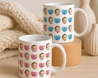 May include: Two white ceramic mugs featuring repeating patterns of a baby's face and heart-shaped graphics. The back mug displays blue hearts with the text I LOVE YOU DADDY, while the front mug features pink hearts with the text I LOVE YOU MUMMY.