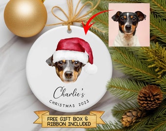 Könnte beinhalten: Weißer Keramik-Weihnachtsbaumschmuck mit einem Hund, der einen roten Weihnachtsmann-Hut trägt. Der Name des Hundes ist "Charlie's" und das Jahr "2023" ist darunter gedruckt. Der Baumschmuck ist von einem grünen Tannenzweig und einem goldenen Band umgeben.