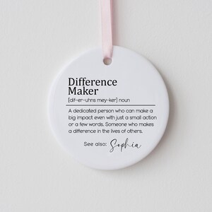Op de afbeelding: Ronde witte ornament met zwarte tekst die luidt: "Difference Maker [dif-er-uhns mey-ker] noun Een toegewijde persoon die zelfs met een kleine actie of een paar woorden een grote impact kan hebben. Iemand die een verschil maakt in het leven van anderen. Zie ook: Sophia"