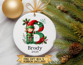 Op de afbeelding: Een witte keramische kerstbal met een feestelijk elf-ontwerp, de letter "B" en de naam "Brody" eronder. De ornament heeft een rood-groen kleurenschema, met sneeuwvlok- en hulstaccenten. Het jaar 2025 staat er ook op.