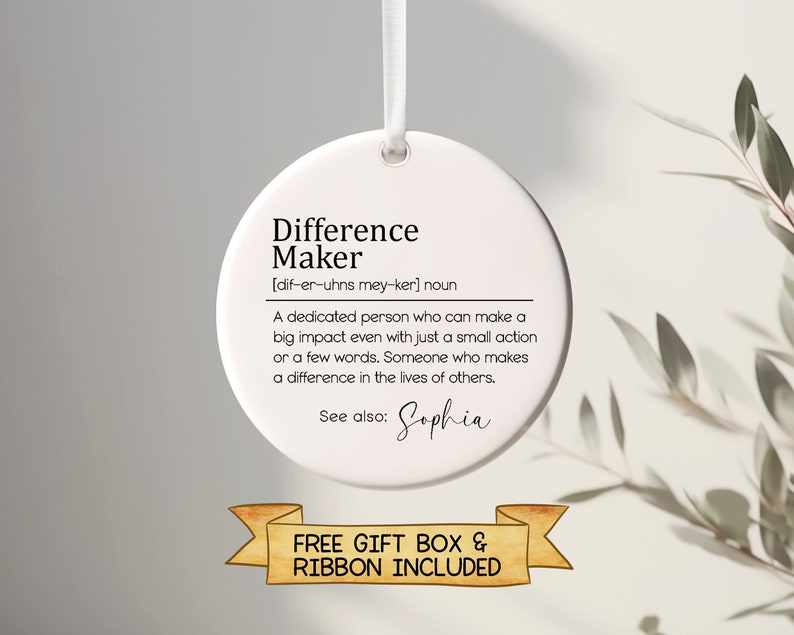 Op de afbeelding: Ronde witte ornament met de tekst "Difference Maker" en een definitie van het woord. De tekst "Zie ook: Sophia" staat onder de definitie. De ornament hangt aan een wit lint. De tekst "FREE GIFT BOX & RIBBON INCLUDED" staat onder de ornament.