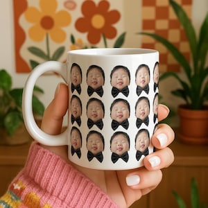 May include: A white ceramic mug featuring a repeating pattern of a child's face with eyes closed and tongue slightly out, wearing a black bow tie. The design is printed in a grid layout across the exterior surface of the cylindrical mug.