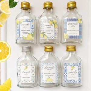 May include: Six small glass bottles with gold and silver caps, each labeled "Limoncello with love." The labels feature lemon illustrations and names with dates. The bottles are arranged on a white surface, with lemon slices in the background.