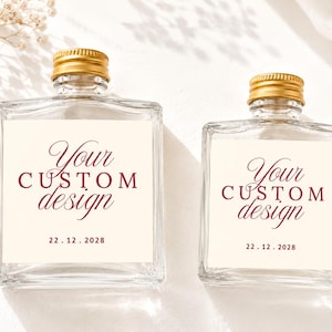 May include: Two clear glass bottles with gold screw-top lids. Each bottle features a square cream-colored label with the maroon text "Your CUSTOM design" and the date "22.12.2028" printed in a combination of script and serif fonts.