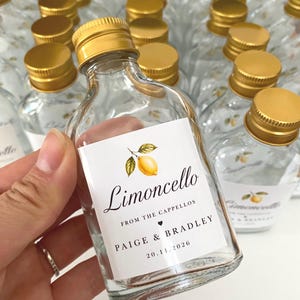 May include: A small clear glass bottle with a gold screw cap, featuring a white rectangular label with a yellow lemon illustration and the text Limoncello, FROM THE CAPPELLOS, PAIGE & BRADLEY, and the date 20.11.2026 printed in black ink.