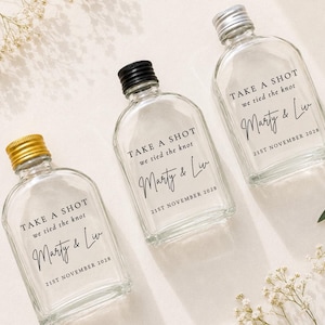 Op de afbeelding: Drie doorzichtige glazen flessen met schroefdoppen in goud, zwart en zilver. Elke fles is voorzien van zwarte tekst: TAKE A SHOT we tied the knot, Marty & Liv, 21ST NOVEMBER 2028. De flessen hebben een afgeronde, rechthoekige vorm.