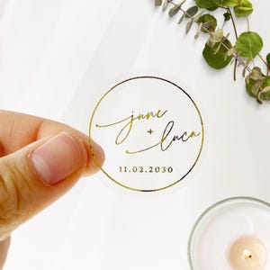 Może przedstawiać: Przezroczysta, okrągła naklejka ze złotym napisem i złotą obwódką. Tekst brzmi "jane + luca" nad datą "11.02.2030". Naklejka trzymana jest w dłoni, idealna na wesela.