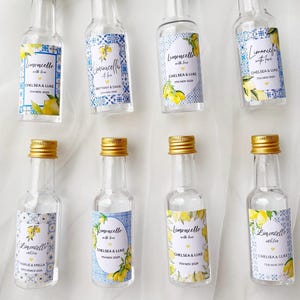 May include: Eight clear glass miniature bottles with gold screw caps, each featuring a custom label with blue and white Mediterranean-style tile patterns, yellow lemon illustrations, and printed text including Limoncello with love.