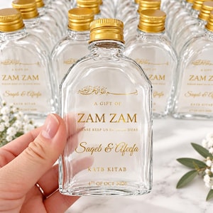 May include: A clear glass bottle with a gold screw cap, featuring gold printed text that reads A GIFT OF ZAM ZAM, PLEASE KEEP US IN YOUR DUAS, Sageb & Afeefa, KATB KITAB, and 4TH OF OCT 2026, along with Arabic calligraphy at the top.