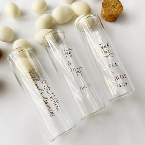 May include: Three clear glass test tubes with cork stoppers, filled with white candy-coated almonds. Each tube features gold foil text: "THANK YOU FOR CELEBRATING WITH US Nathaniel & Jasmine", "Matt & Nat 28/06/29", and "Thank You SOFIA & GEORGE 20.04.29".
