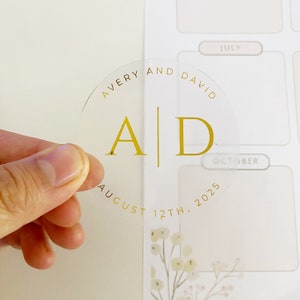 Könnte beinhalten: Ein runder, transparenter Aufkleber mit goldfarbenem Foliendruck. Das Design zeigt oben die Namen AVERY AND DAVID, in der Mitte die Initialen A | D und unten das Datum AUGUST 12TH, 2025, alles in einer klaren Serifenschrift gedruckt.