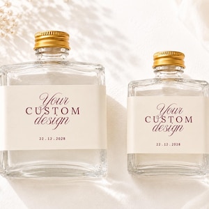 May include: Two clear glass bottles of different sizes with gold screw caps. Each bottle features a cream-coloured rectangular label with the dark red text "Your CUSTOM design" and the date "22.12.2028" printed in a clean, centred layout.
