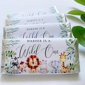 May include: Chocolate bar wrappers with a jungle theme. The wrappers feature the text "HARPER IS A Wild One" in elegant script, along with illustrations of a zebra, monkey, lion, hippo, elephant, and giraffe. Green foliage accents the design.