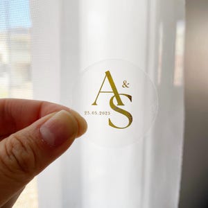 Può includere: Un adesivo rotondo trasparente con scritte dorate. Il design presenta le iniziali "A&S" e la data "25.05.2025". L'adesivo è tenuto su uno sfondo bianco, suggerendo un tema di matrimonio o evento.