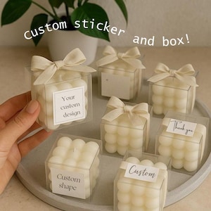 Könnte beinhalten: Mehrere quadratische, blasenartige Kerzen in transparenten Boxen, gebunden mit beigen Schleifen. Jede Box hat ein Etikett mit Text wie "Your custom design" und "Custom shape". Das Bild enthält auch den Text "Custom sticker and box!"
