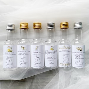 May include: Six small glass bottles with silver and gold caps. Each bottle has a white label with text and olive branch illustrations. The labels read "Thank You" and names, with the date "7th OCT 2026".