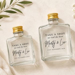 May include: Two clear glass bottles with gold screw caps, each featuring the black printed text: TAKE A SHOT we tied the knot, Marty & Liv, 21ST NOVEMBER 2028. The bottles are square-shaped with rounded corners and a short neck, designed for liquids.