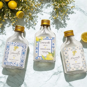 May include: Three small glass bottles of Limoncello with gold caps and decorative labels. The labels feature a lemon illustration and text, with dates and names. The bottles are arranged on a marble surface with lemons and flowers.