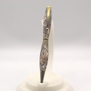 May include: A decorative pen with a floral pattern in shades of brown, white, and gray. The pen has a gold-colored clip and tip. The pen is displayed on a white stand.
