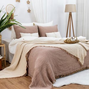 Puede incluir: Una manta de tiro de color marrón claro y beige con flecos en los bordes sobre una cama con ropa de cama blanca y almohadas marrones. La manta está hecha de un tejido suave y texturizado.