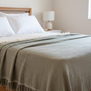 Puede incluir: Una cama está hecha con sábanas y almohadas blancas, cubierta con una manta verde salvia. La manta tiene un tejido texturizado y flecos en el borde inferior. También se ve un marco de cama de madera y una lámpara de noche blanca.