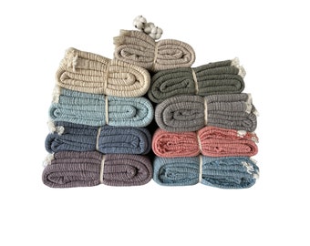 May include: A stack of eight folded cotton throws in various colors, including light blue, gray, pink, and beige. The throws are arranged in a graduated color scheme, with the lightest color at the top and the darkest color at the bottom.