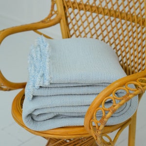 Peut inclure: Un plaid bleu clair avec une bordure frangée est plié et placé sur une chaise en osier. La chaise a un dossier tissé et un siège rond.