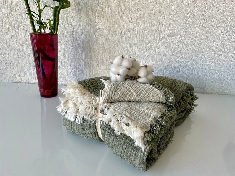 May include: A green and white waffle weave throw blanket with fringe trim. The blanket is folded and tied with a white ribbon. Cotton bolls are placed on top of the blanket.