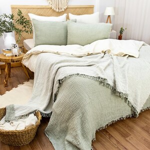 May include: A sage green waffle weave throw blanket with fringe edges is draped over a bed with white and sage green bedding. The bed has a wooden headboard with a woven pattern.