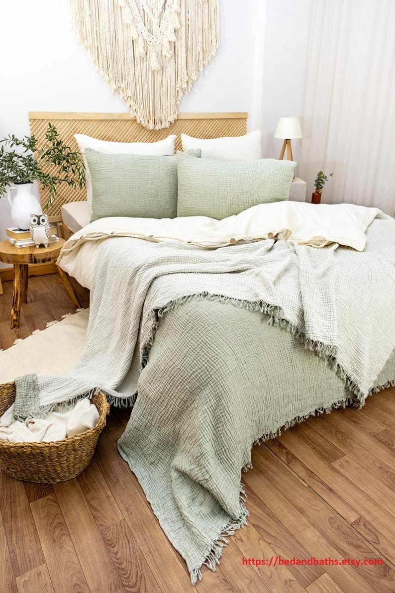 May include: A sage green waffle weave throw blanket with fringe edges is draped over a bed with white and sage green bedding. The bed has a wooden headboard with a woven pattern.