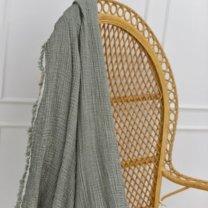 May include: A sage green throw blanket with a fringed edge draped over a wicker chair. The blanket is made of a soft, textured fabric.