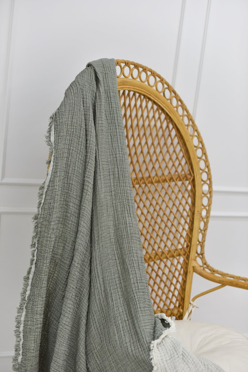 May include: A sage green throw blanket with a fringed edge draped over a wicker chair. The blanket is made of a soft, textured fabric.