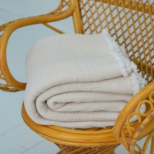 Puede incluir: Una manta de tejido de waffle blanco con un borde de flecos está doblada y colocada en una silla de mimbre. La silla tiene un respaldo tejido y una base redonda.