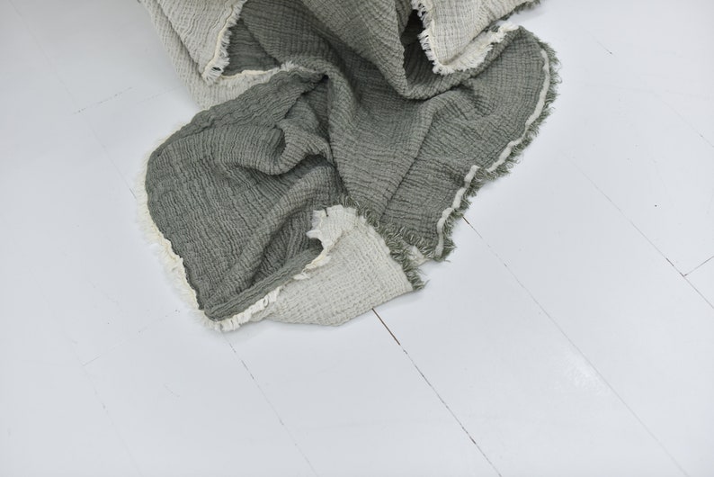 May include: A soft, green and white throw blanket with a fringed edge. The blanket is made of a waffle weave fabric and is folded on a white wooden floor.