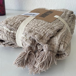 May include: A beige throw blanket with tassels and a woven texture. The blanket is folded and tied with a white ribbon. The label on the blanket reads "The Morrow".