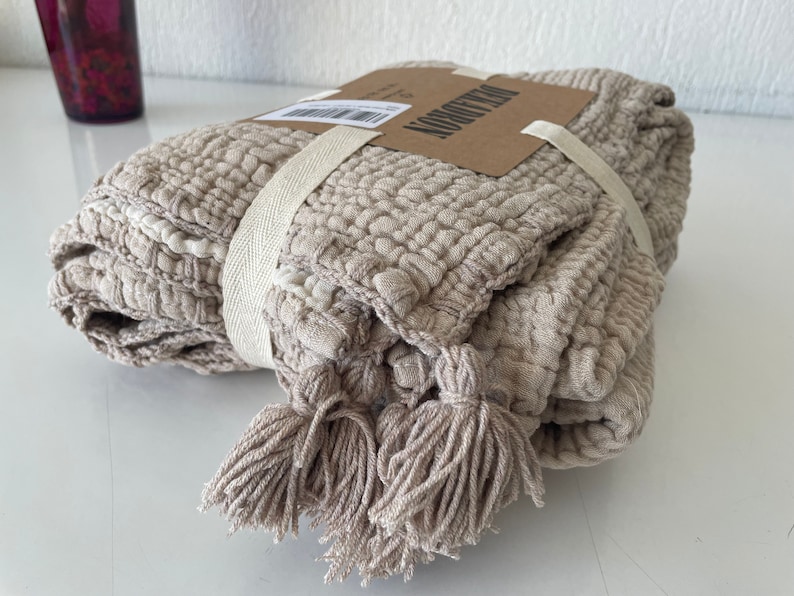 May include: A beige throw blanket with tassels and a woven texture. The blanket is folded and tied with a white ribbon. The label on the blanket reads "The Morrow".