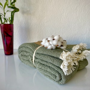 May include: A folded sage green throw blanket with a white cotton flower accent and a brown ribbon tied around it. The blanket has a fringe border.