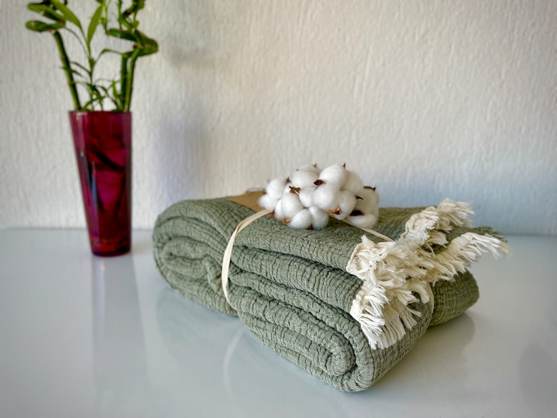 May include: A folded sage green throw blanket with a white cotton flower accent and a brown ribbon tied around it. The blanket has a fringe border.