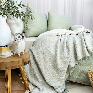 May include: A green and white waffle weave throw blanket with fringe edges. The blanket is draped over a bed with green and white pillows. A white ceramic owl figurine sits on a wooden table next to a vase of green plants.