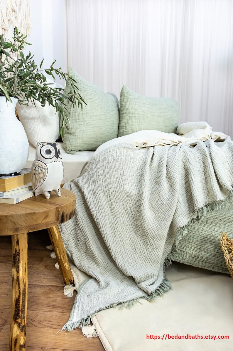 May include: A green and white waffle weave throw blanket with fringe edges. The blanket is draped over a bed with green and white pillows. A white ceramic owl figurine sits on a wooden table next to a vase of green plants.