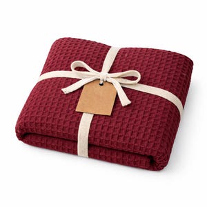 May include: A folded burgundy waffle-weave blanket tied with a cream-colored ribbon and bow. A blank brown tag hangs from the ribbon. The blanket is a solid color and has a textured surface.