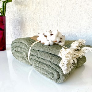 May include: A sage green throw blanket with a textured weave and fringe trim. The blanket is folded and tied with a natural twine ribbon. A bunch of white cotton bolls are placed on top of the blanket.