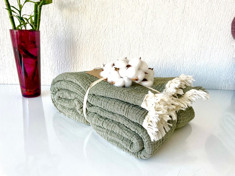 May include: A sage green throw blanket with a textured weave and fringe trim. The blanket is folded and tied with a natural twine ribbon. A bunch of white cotton bolls are placed on top of the blanket.