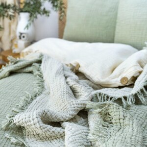 May include: A soft, textured throw blanket in a light green and white color scheme. The blanket has a fringed edge and is folded on a bed with matching pillows.