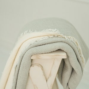 Peut inclure: Une couverture douce en coton de couleur vert clair et blanc avec une bordure frangée, pliée et drapée sur un tabouret en bois blanc.