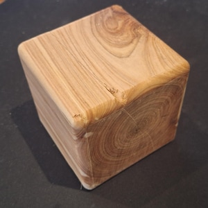 May include: A solid wooden cube featuring a natural light brown grain pattern with visible growth rings on the top surface. The block has smooth, rounded edges and a distinct crack running across the top corner, showcasing the raw texture of wood.