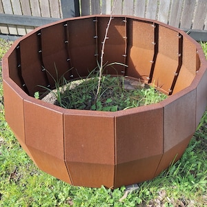 May include: A large, rusty brown, metal planter with a rounded, octagonal shape. The planter is made of multiple panels that are joined together with screws. The planter is sitting on a bed of green grass.